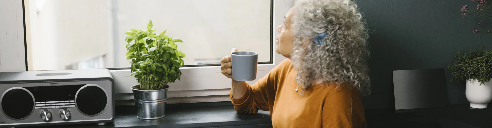 Woman with audio processor holding a mug by a window near a radio and plants