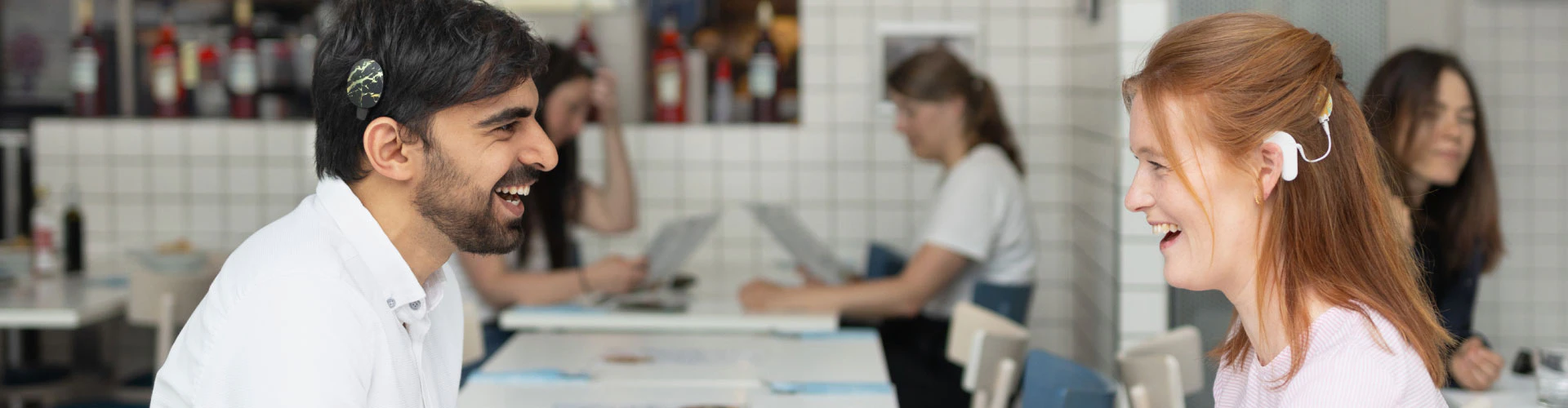 A man and a woman, both with hearing devices, talking in busy café setting