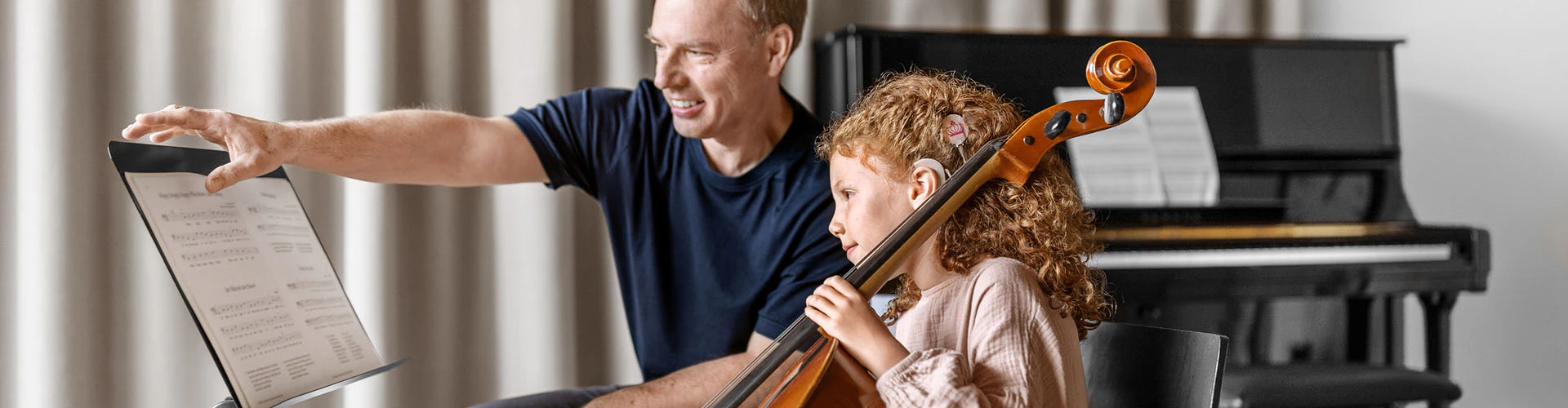 A child with a cochlear implant practices cello beside an instructor who points toward a sheet‑music stand. There are a piano and curtains in the background.