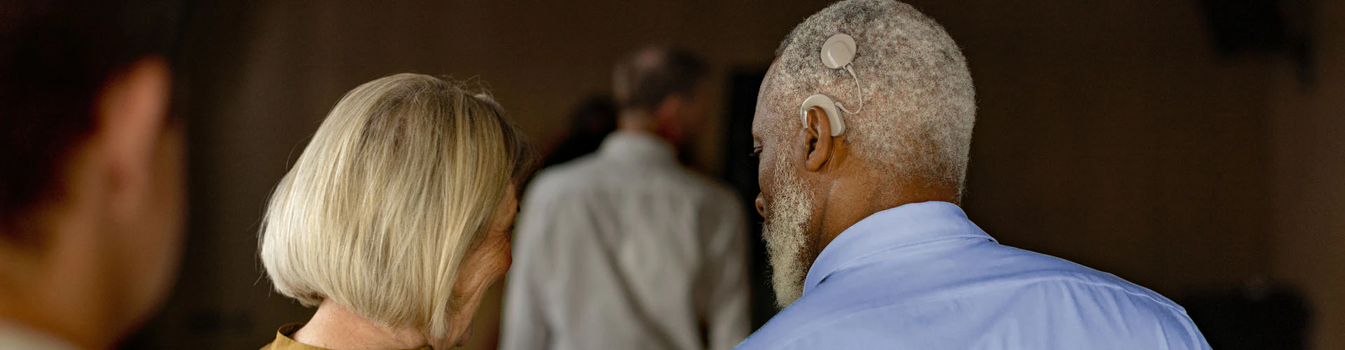 Two people walking side by side in a softly light corridor. The man has a cochlear implant audio processor visible on behind his ear.