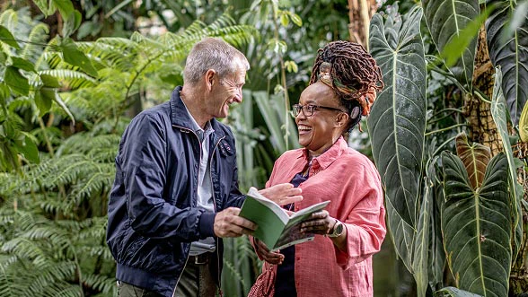 A man and a woman, who is wearing a CI audio processor, are standing in a botanical garden, looking at a book