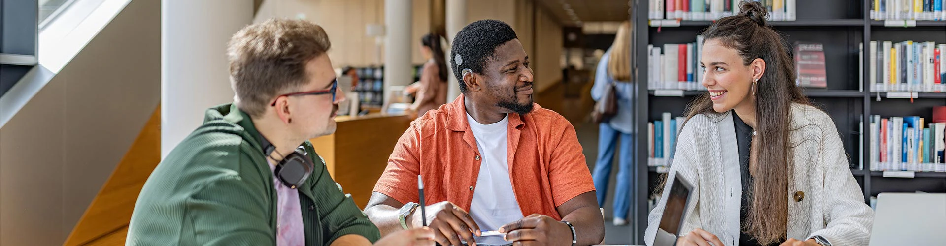 2 men, one man with a cochlear implant, and a woman sitting in the library