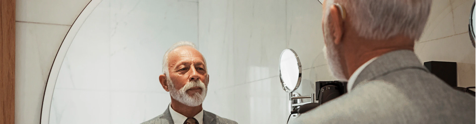 A man with a hearing aid looks into a large mirror in a bathroom