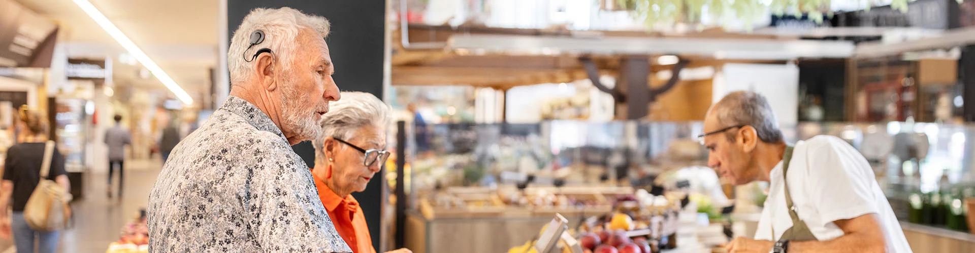 Two older people at a market counter. The man has a cochlear implant audio processor