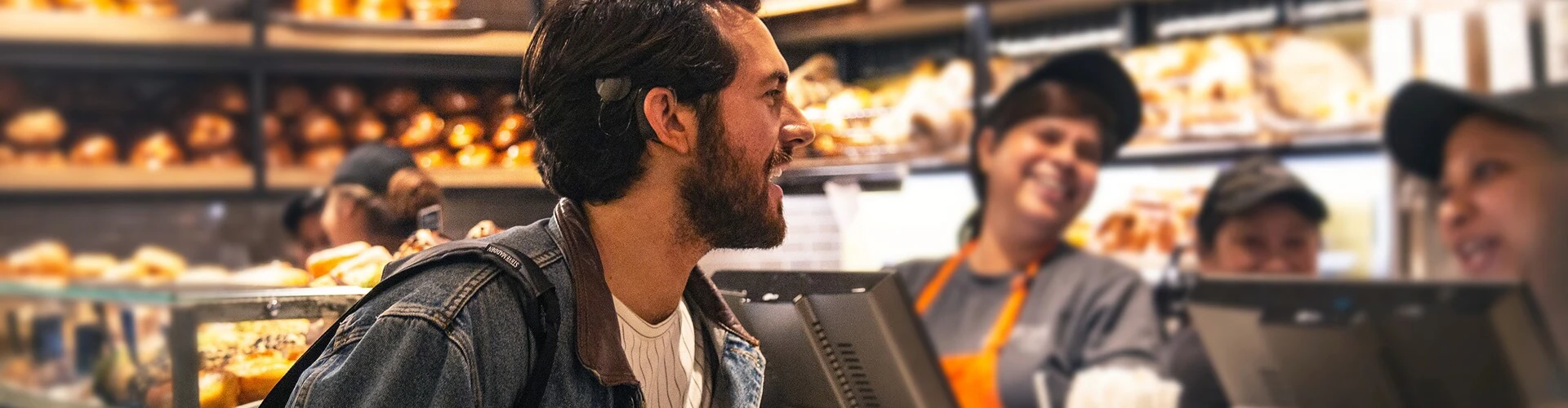 Man at a bakery counter wearing a SONNET 2 behind the ear, interacting with staff.