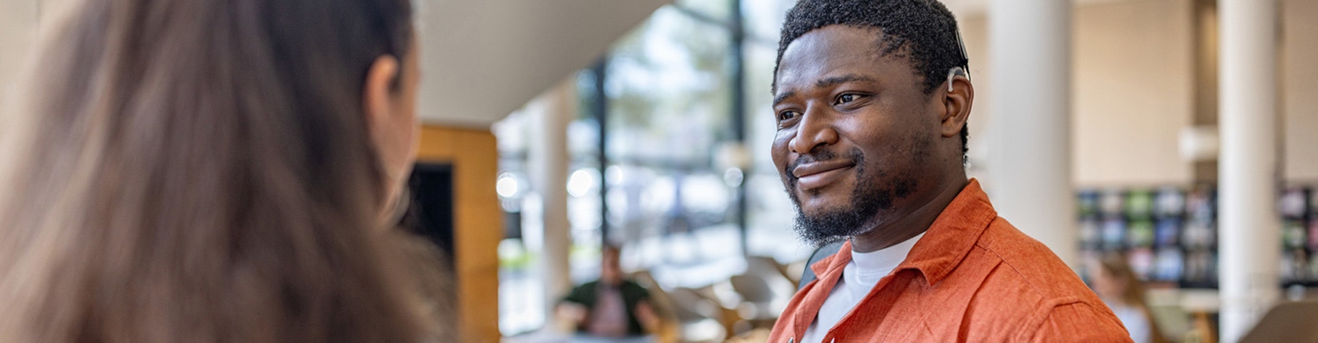 A man and a woman talk in a bright indoor setting. The man is wearing a cochlear implant audio processor behind his ear.