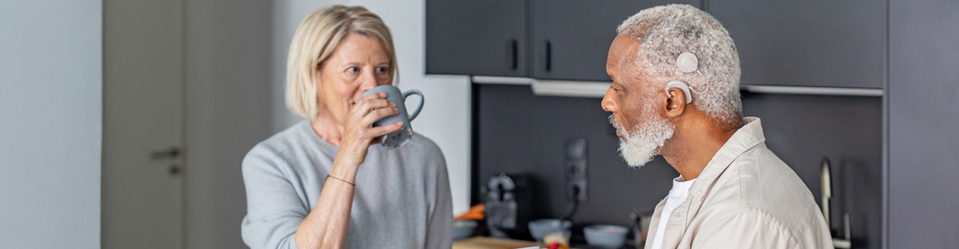 A retired couple are talking in a kitchen. The man wears a cochlear implant audio processor behind the ear.