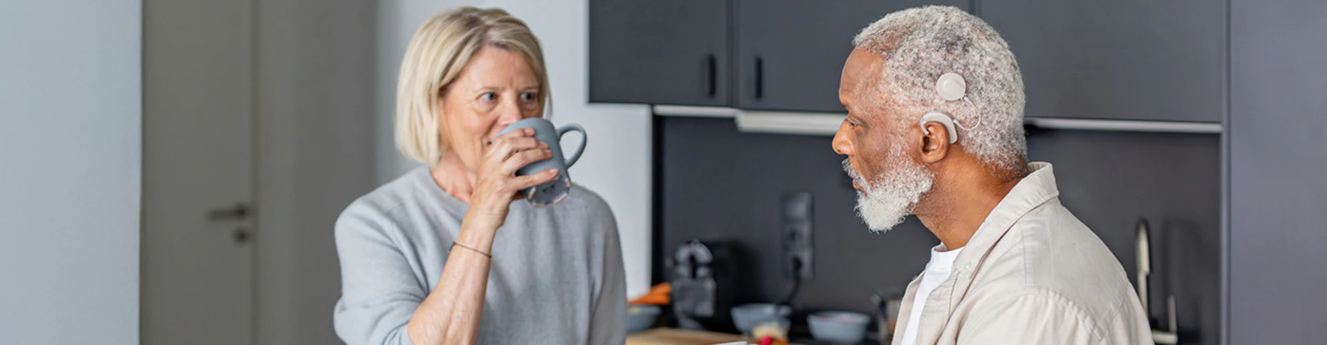 A retired couple are talking in a kitchen. The man wears a cochlear implant audio processor behind the ear.