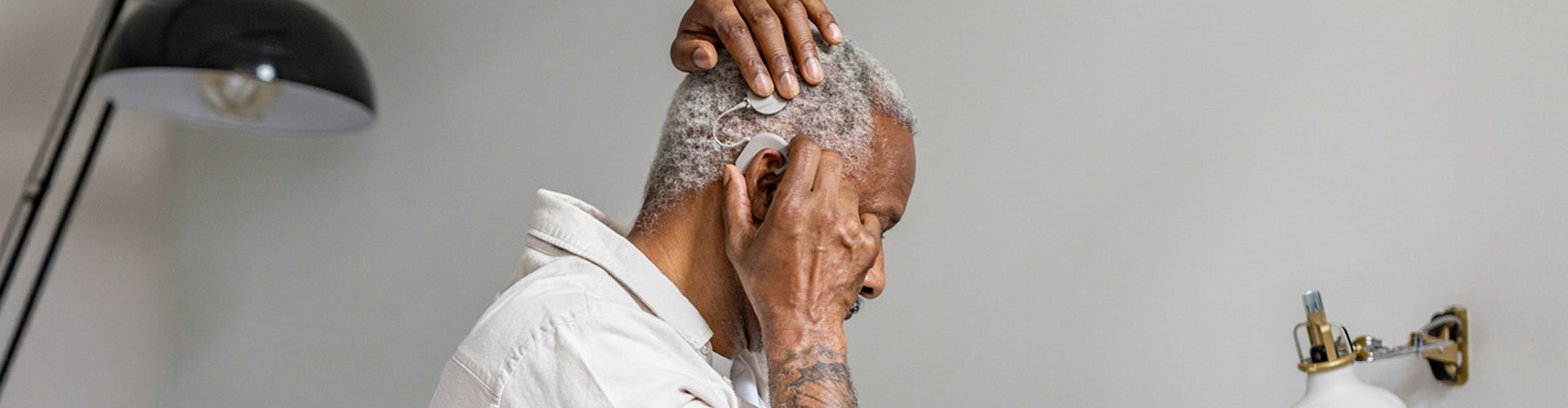 An older man puts on his SONNET 3 cochlear implant audio processor while indoors near a lamp.