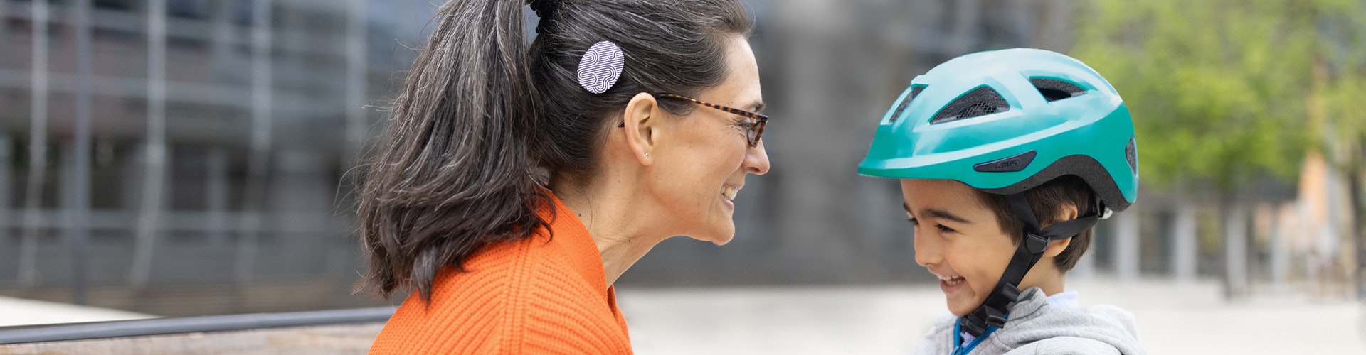 A mother wearing a RONDO 3 cochlear implant audio processor smiles at her young son, who is wearing a green bike helmet.