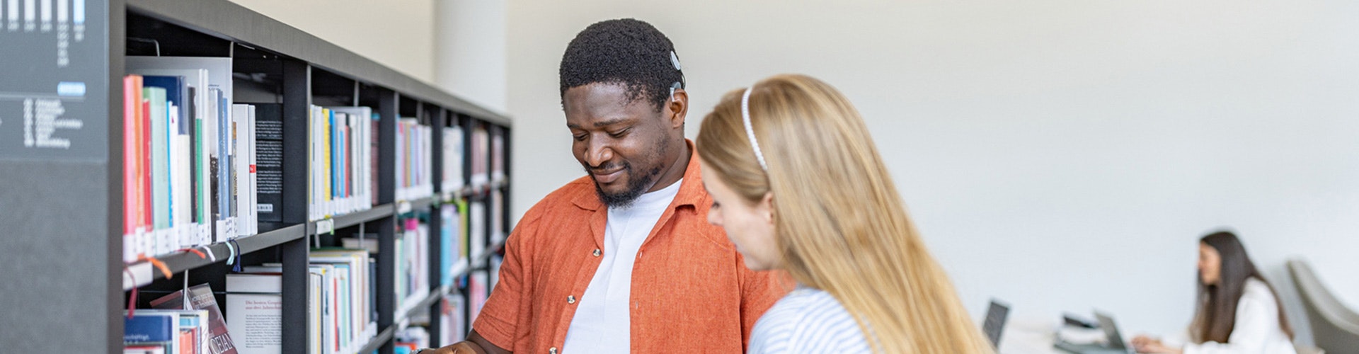 Two people stand in front of a bookshelf in a library. One person is wearing a cochlear implant audio processor behind his ear.