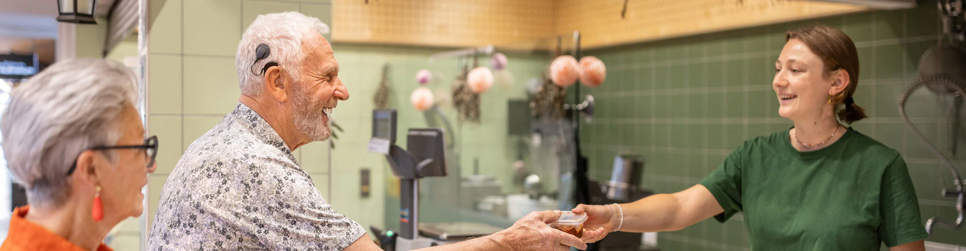 A couple stand by a shop counter. The person with an audio processor receives an item