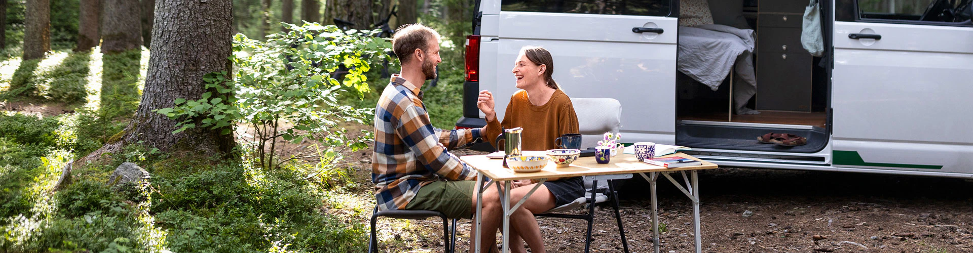 Two people sitting outside a camper van in a forest campsite.